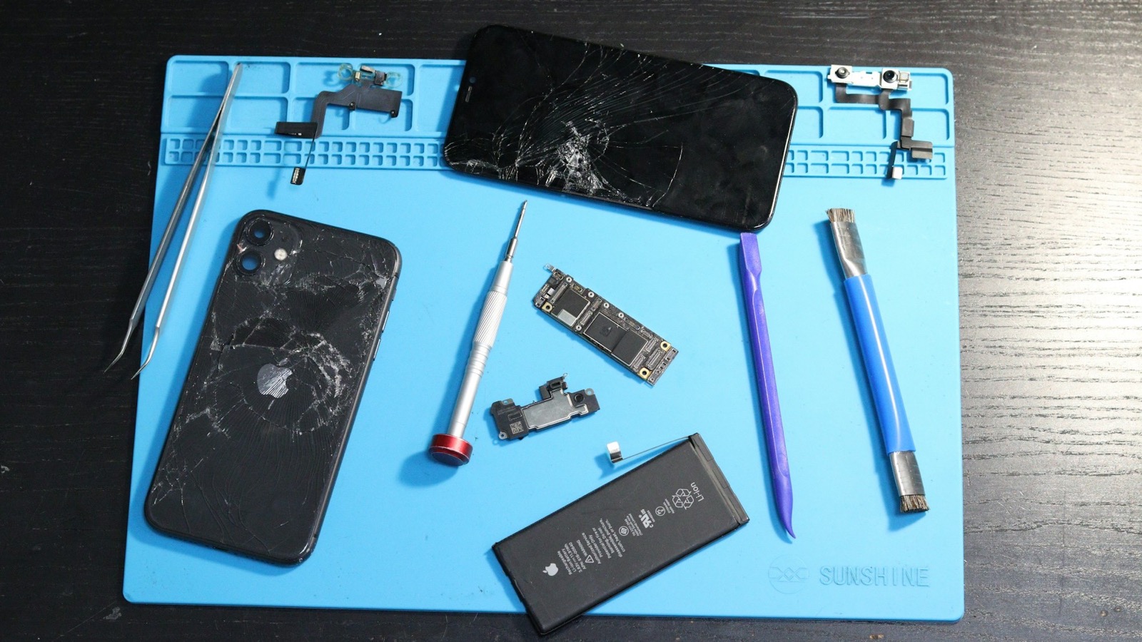 Disassembled iPhone with repair tools, logic board and battery laid out on a technician's repair mat
